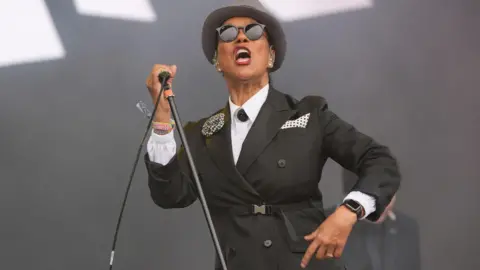 Pauline Black of The Selecter is on stage in a fitted black suit jacket and white shirt. She is holding a microphone stand as she sings.