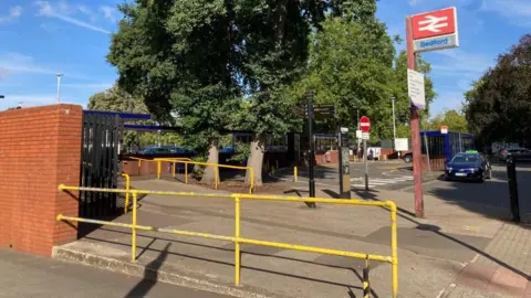 Alex Pope/BBC Outside Bedford railway station, showing yellow railings, a sign for Bedford Station, a bus stop in the distance, a parked taxi and several trees. 