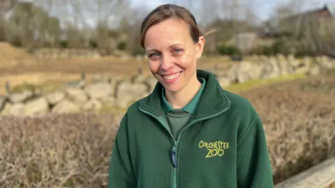 Qays Nahim/BBC Rebecca Moore, zoological director of Colchester Zoo, looking at the camera and smiling. She is wearing a green polo shirt, jumper and fleece jacket with yellow Colchester Zoo branding on her left chest. She is standing in front of an outdoor animal enclosure.