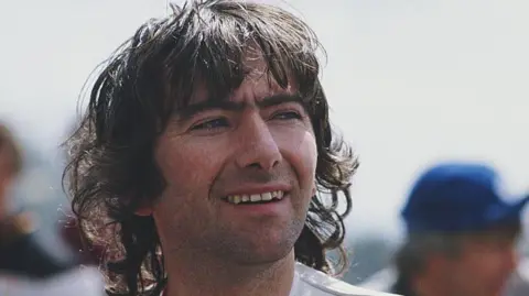 Getty Images Joey Dunlop has long dark brown hair with a fringe, he has dark eyebrows and facial stubble. He is wearing a white racing suit and is looking to his left, the background is blurred.