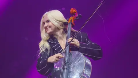 Getty Images A woman with long blonde hair is on a stage. She is holding a silver glittery cello and a bow. The light behind her is purple