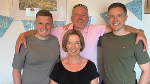 Kate Turner Kate surrounded by her husband and two adult sons, who have their arms around each other and they are all smiling to camera. There is blue bunting on the wall behind saying '30'