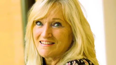 Karen Loweman Karen Loweman has long blonde hair and is wearing a floral dress. She is smiling at the camera indoors.