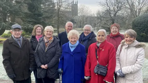 Whittle Productions Nine people in a park. Seven women and two men. They are all people who contributed to the memories of the lights festival project. They are all wearing coats and scarves and the ground behind is frozen.