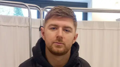 Stephen Huntley/BBC A head and shoulders image of Callum Reid. He is wearing a black hooded top and looking directly into the camera. Behind him is a white curtain hanging from a curtain rail similar to those seen in hospitals.