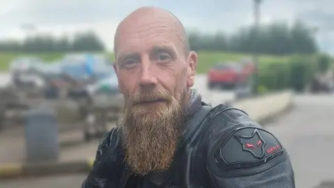 A man with a bald head and long beard looking ahead. He is wearing a black leather, biker-style jacket.