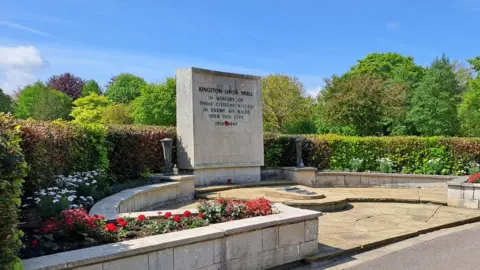 A stone memorial area. The memorial is surrounded by leafy green trees and hedges with red and white flowers planted in front of a walled area. An inscription on the memorial reads "In memory of those citizens killed in enemy air raids over this city".