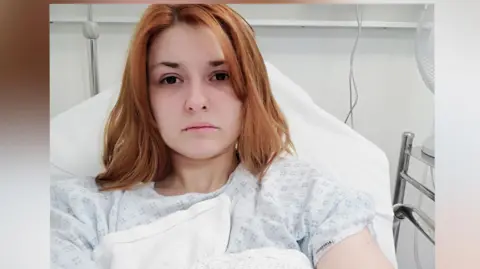 A woman sat in a hospital bed staring at the camera, she has ginger hair. 