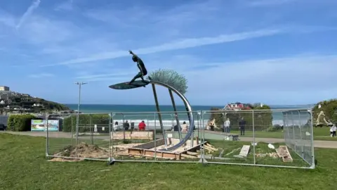 The bronze statue of a surfer behind metal fencing, in its position overlooking a beach and some people walking behind.