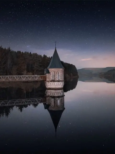 Réservoir Cormac Downes Pontsticill à Powys avec la tour de vannes et le pont au-dessus de l'eau photographiés sur fond de ciel étoilé. La tour et le pont se reflètent parfaitement dans l'eau en contrebas et il y a des arbres et des montagnes en arrière-plan.