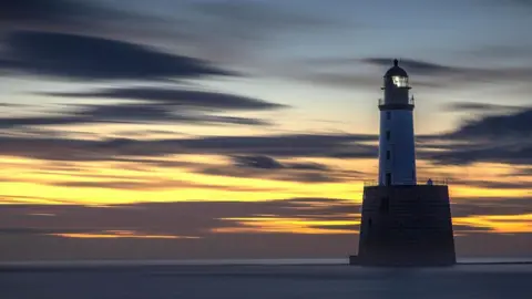 A lighthouse in the sea. The sky is orange with dark clouds streaked across it.