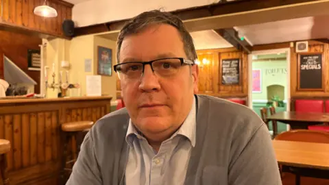 Martin Heath/BBC Simon Clawson with short brown hair and glasses looking at the camera wearing a beige pullover and light-coloured shirt. He is sitting in a pub with the bar to his right and specials boards behind him.