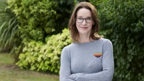 Hay Festival A woman with shoulder-length brown hair and glasses, wearing a light blue/grey jumper stands with her arms folded in front of trees and bushes, smiling at the camera.