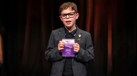 A young boy with short brown hair and wearing glasses and a dark grey suit. He is holding a purple and white award in both hands. Behind him is a dark red or brown curtain. 