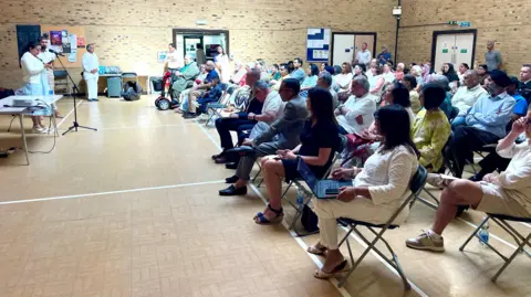 Stephen Huntley/BBC People sit in a community centre on chairs that look toward a microphone. A woman is standing behind the microphone reading from a phone in her hands. Two men stand close to her as she reads. The picture is taken from the side of the hall.