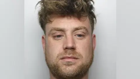 Close crop of a police mugshot. The man has a wide lower face and large eyes. He is looking down, slightly past the camera. He has a light brown beard.