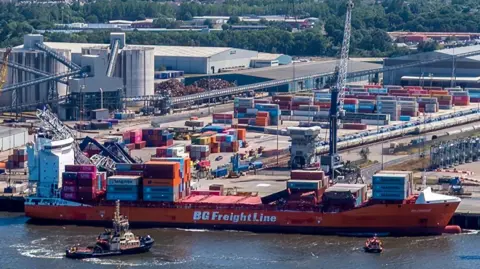 Urbex Drone View Aerial view of the side of a large container ship. It is orange with BG Freight Line on the side, and a number of containers on deck. Behind it and to the left can be seen part of a collapsed crane. Large numbers of containers are stacked on the dock beside it, and on the river next to it is a tug.