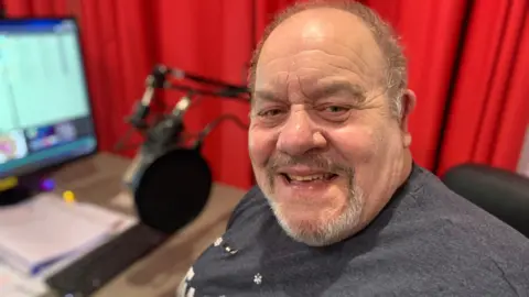 A man with short grey hair and a grey beard is smiling at the camera. He is wearing a grey t-shirt and sitting in front of a microphone and computer