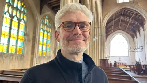 Immy Lee/BBC Jonathan Dowman has white short hair, swept back and is wearing a navy fleece and a blue shirt. He is standing in the rows of pews and in front of the stained glass windows.