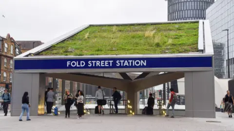 Alamy Passengers entering and exiting the entrance of a tube station with a large blue sign above the archway. The sign has the words "Fold Street tube station".