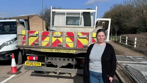 Kirsty Knight Kirsty, a woman, wearing a grey T-shirt and a black cardigan, standing in front of a roadworks truck on the road. 
