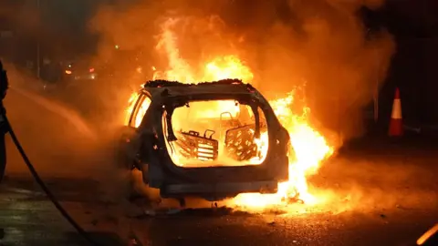 Burning car in the night. There is fire all around and the car is charred with the back completely gone and fire engulfing the inside and the seats. 
