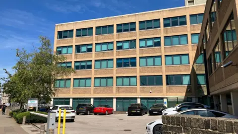 LDRS A five-story office building with a car park in front of it. It is built at a right angle with one block running to the left and the other on the far right. There is a entrance with a barrier across it to the car park, which has a few cars. On the far left is a pavement. 