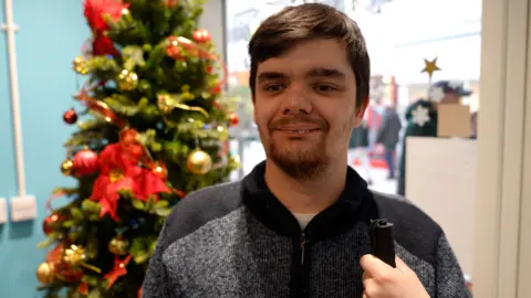 A man with brown hair is wearing a dark zip-up sweater and stands indoors near a decorated Christmas tree with red poinsettias and gold ornaments.