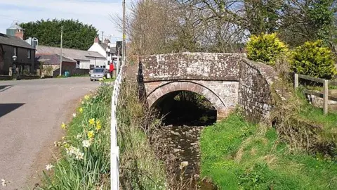 A small stone bridge standing over a small waterway called Pen Beck. Stones can be seen through its shallow water. A road stands to the left of the beck. Village houses and a pub can be seen in the background.