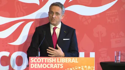 A man with short grey hair and wearing a dark blue suit, white shirt and pink tie is standing behind a lectern on a stage with a sign saying Scottish Liberal Democrats