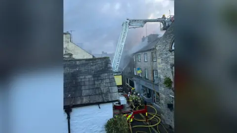 Eugene Kaufman Firefighters on the street in front of the building that caught fire in Kirkby Lonsdale. They are working with a firefighter who is on an aerial ladder to bring the flames under control.