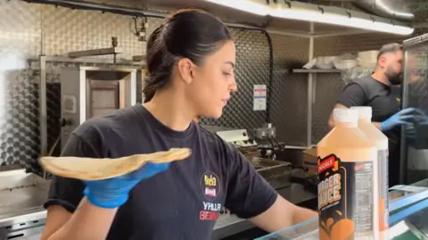 Andrew Sinclair/BBC Sahar in a black t-shirt is working inside her food van. She holds an open wrap in one hand while preparing to fill it. Her husband also in a black t-shirt and with a beard can be seen in the background looking in the fridge. 