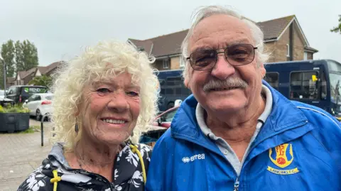 An elderly husband and wife stand in a high street. They are smiling. She has white curly hair and he has a jacket of his local football team on. 