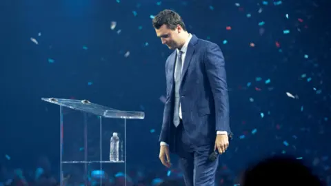 Reuters Charlie Kirk stands in profile with confetti falling around him wearing a blue pinstripe suit on a darkened stage