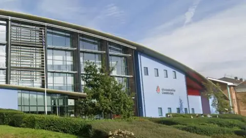 The Gloucestershire Police headquarters, a large blue building with a sloped roof and large windows. At the front of the building, there are lots of hedgerows.