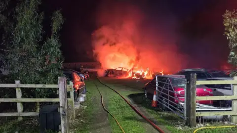 Yard with farm gate with several cares on fire in the yard