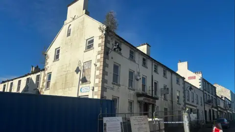 The Antrim Arms Hotel, a white building. Antrim Arms is written in red letters above the door. There are bollards and a fence in front of it.
