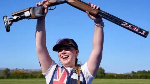 Bethany Norton wears a blue vest with a British flag on one side. She has a white T-shirt on underneath. She is also wearing a blue baseball style cap and is holding the gun above her head in triumph.