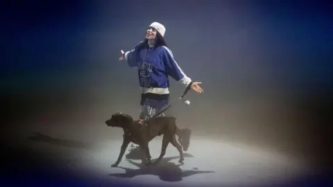 Getty Images Billie Eilish on stage for a listening party for her album Hit Me Hard and Soft in LA in May. Billie is a 22-year-old woman with long dark hair. She wears a white cap twisted backwards and a blue sports shirt. She's on stage with her dog, Shark. 