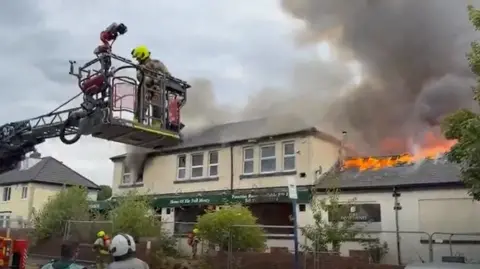 A firefighter on a fire ladder tackling a fire coming from the roof of a two-storey building. Smoke is pillowing into the air.