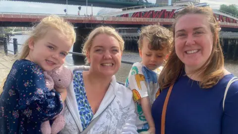 Julie Smith/BBC  Kirsten Ormond and Sarah Walker are with their children. One has a daughter, who is clutching a teddy bear, and the other has a son who is wearing a shirt with wild animals on it. They are all smiling on Newcastle's quayside.