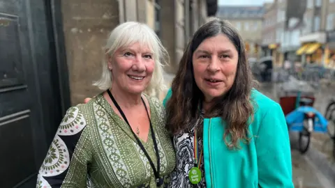 Two women standing next to each other. The one on the left has shoulder-length white hair and is wearing a green top and a black lanyard. The woman on the right has long brunette hair and is wearing a green jacket over a white and black top.