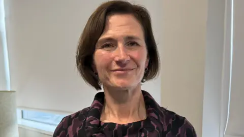 Annette Morris has short, brown hair wearing a dark purple and black patterned dress with a cowl neck in an office setting.