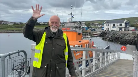 Derek Adams with short white hair and beard, wearing a dark green waterproof coat and yellow hi-vis jacket. He is waving with his right hand. Behind him is the orange RNLI Charles Lidbury lifeboat, moored alongside a landing stage with white railings. The sea is visible in the background.