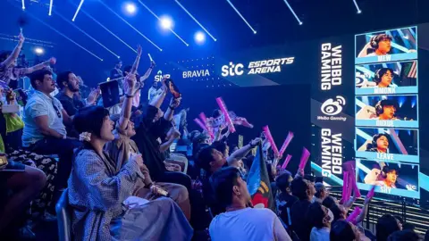 Getty Images A section of the crowd celebrates as they watch a play during the esports world cup. Foam fingers point into the air and eyes are directed towards a tower of screens showing the faces of individual players.