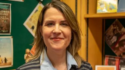 Joanne in a library wearing a black and white striped suit jacket and light blue collared shirt. She has fair hair to her shoulders.