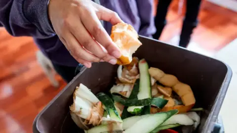 A black box containing food waste can be seen with a person about to drop an apple core in. They are standing in a room with a hard brown wooden floor.