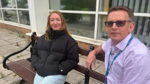 Lowri and Dr Healy sitting on a bench. Dr Healy is wearing a purple shirt with a lanyard, he has short dark hair and wears glasses. Lowri is wearing a black puffer jacket and jeans. 