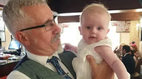 Caroline Thwaites Colin Thwaites is pictured at a function with a grandchild. Colin is wearing glasses and a Harris Tweed waist coat over a white shirt and a dark blue tie.