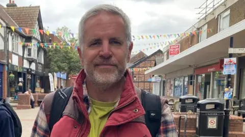 Paul Esling, a resident of Northwich, in the town centre. He has short grey hair, a trimmed beard, and wears a lime green t-shirt with a plaid shirt and red jacket.